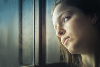 A teenager girl looking away through a window at home, thinking, sad.