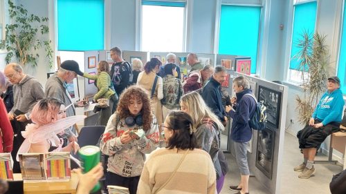 A room of 30 people eat cake, look at art displayed on boards in the reception area of BHT Sussex.